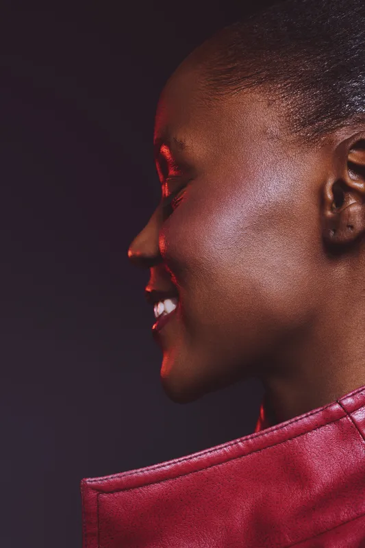 Profile of smiling African woman with dark skin in red leather jacket against dark background. Concept of happiness, fashion, beauty, and confident female portrait in studio lighting.
