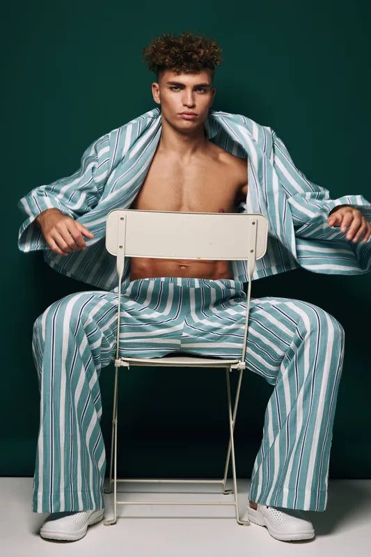 Young man sitting on chair wearing oversized striped suit with open jacket revealing bare chest in studio with dark green background fashion posing for portrait.