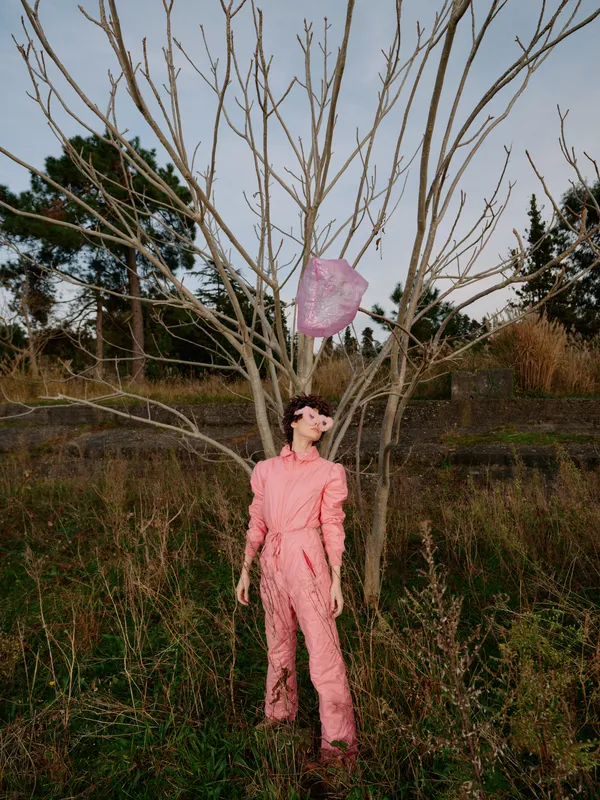 Female in a pink jumpsuit stands beside a bare tree in an open field, wind catching a scarf, creating a dramatic fashion moment in natural daylight and rustic landscape.