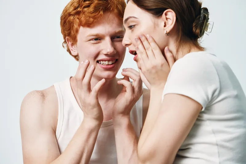 Young couple, man and woman whispering secrets with close expressions and playful mood on white background. Friends sharing private talk and smiling in casual clothing for fun moments together.