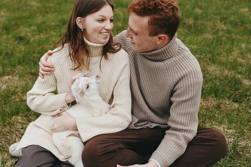 Couple outdoors on grass wearing sweaters, sharing affection with a small dog, romance and companionship in a relaxed lifestyle moment.