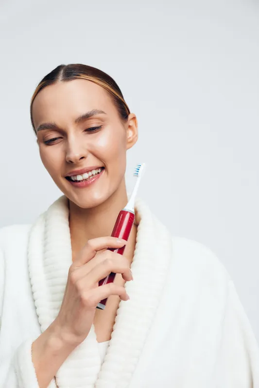 Young woman in white bathrobe smiles happily holding red electric toothbrush close to face isolated on white background with closed eyes and fresh skin.