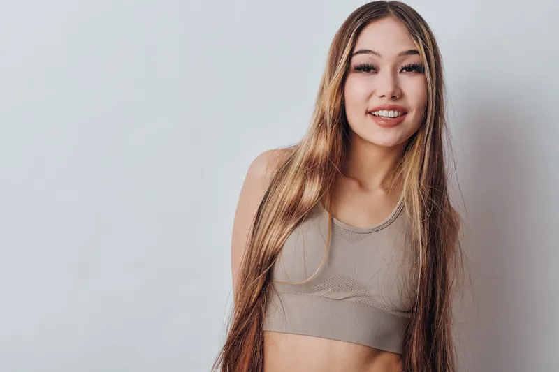 Young woman with long hair and casual top smiling and posing on gray background. Female model with natural makeup and relaxed expression looking at camera indoors.