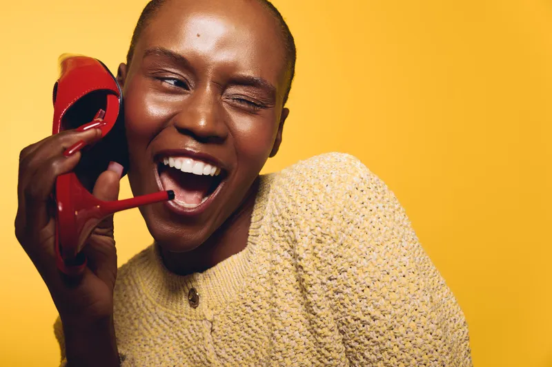 Happy African woman with dark skin wearing yellow sweater holds red high heel shoe near ear as phone, winking and smiling on yellow background. Concept of joy, fashion, and creativity.