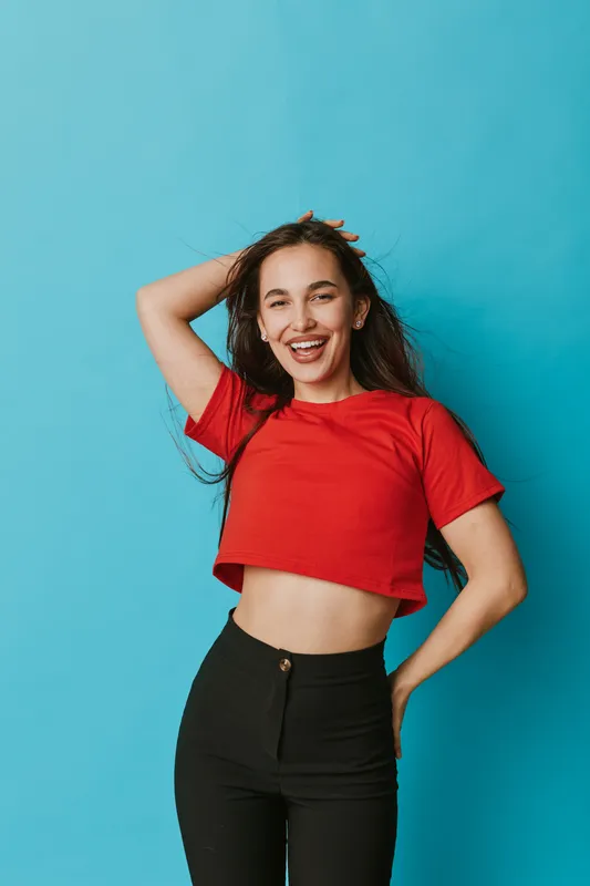 Young woman smiling with hand in hair wearing red crop top and black pants standing against blue background. Casual style, confident pose, happy expression, lifestyle and fashion portrait.