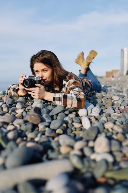 Female photographer lying on a pebble beach with a camera, capturing candid coastal moments in a sunny outdoor scene with a focused expression and creative framing