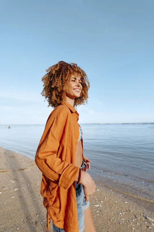 A young woman with curly hair wearing an orange shirt stands on a sandy beach near calm water under a clear blue sky. She looks happy and relaxed enjoying nature.