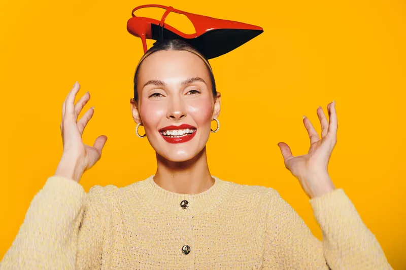 Happy woman wearing a small red and black hat smiles broadly with raised hands on bright yellow background. Concept of joy, fashion, and positive emotions in studio portrait.