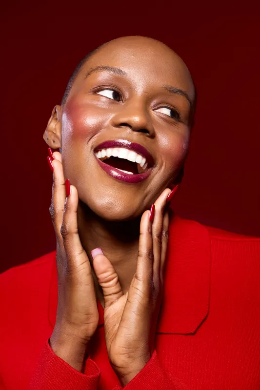 Smiling black woman with dark skin wearing red jacket poses with hands near face on red background. Concept of joy, fashion, beauty, and confident female model with natural makeup.
