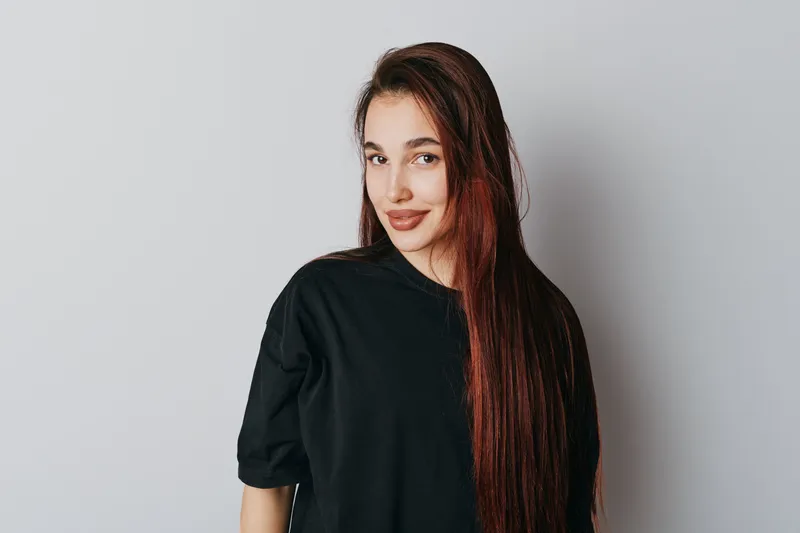 Young woman with long hair wearing black shirt standing against white background looking at camera with slight smile in casual portrait style. Female model with natural makeup and relaxed posture.