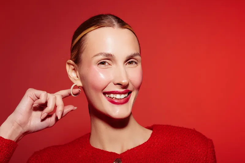 Smiling woman with red lipstick and red outfit poses against red background. Close-up portrait with hoop earrings and slicked back hair. Concept of beauty, fashion, and confidence.