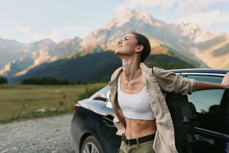 A woman stands beside an open car door on a sunny mountainside, arms outstretched, wearing a white top and beige jacket, enjoying freedom and nature in a relaxed lifestyle scene.