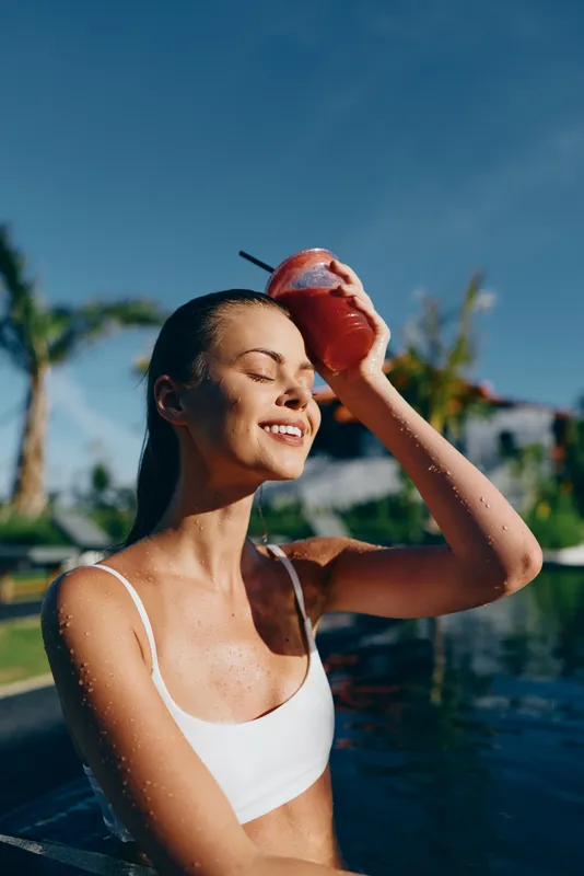 Smiling woman in a white bikini by a sunny pool, enjoying an outdoor skincare moment. Bright blue sky, lush background, relaxed vibe, warm breeze, wellness and self care in a summery scene.