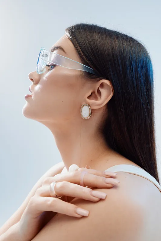 Profile of a young woman with long dark hair wearing transparent glasses and elegant earrings. Soft lighting highlights her smooth skin and natural beauty in a minimalistic setting.