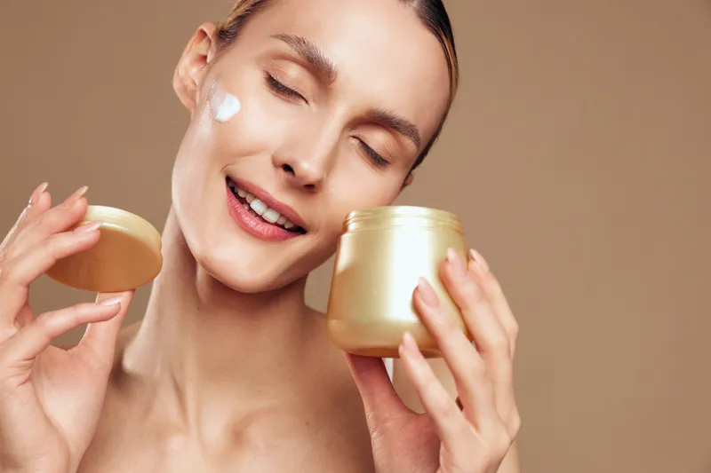 Smiling woman with minimal makeup holds gold skincare jars, applying cream to her cheek. Close-up on a warm beige background. Concept of skincare, wellness, and selfcare. Soft lighting creates a warm