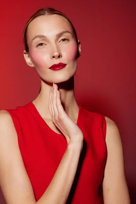 Elegant woman with red lipstick and sleeveless dress poses with hand near face on red background. Studio portrait with soft lighting highlights beauty and fashion concept.