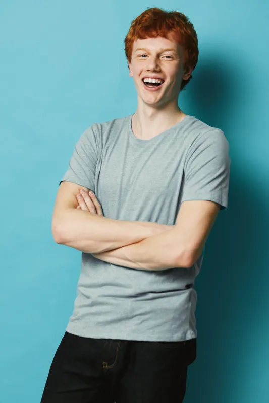 Young man with red hair smiling and laughing with arms crossed wearing gray t shirt and black pants standing against blue background in studio portrait.
