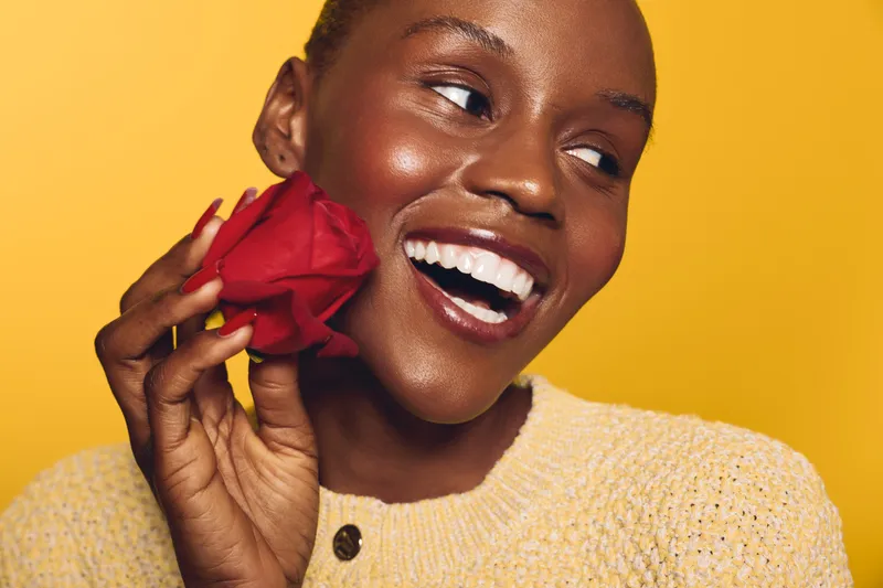 Happy dark skinned African woman smiles joyfully holding red rose near face. Close-up portrait on yellow background. Concept of beauty, happiness, floral, and natural skin glow.