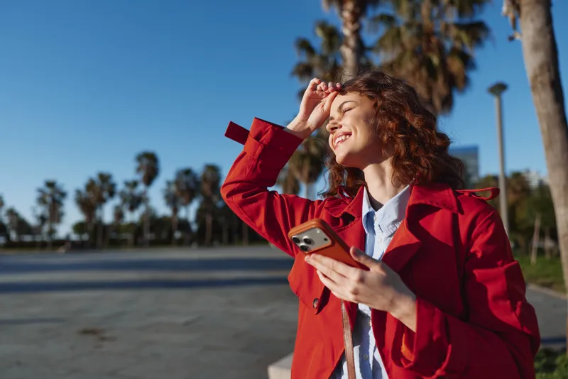 A smiling young woman in a red coat holds a smartphone outdoors along a palm tree lined street, basking in bright sunlight with a carefree, fashionable mood.