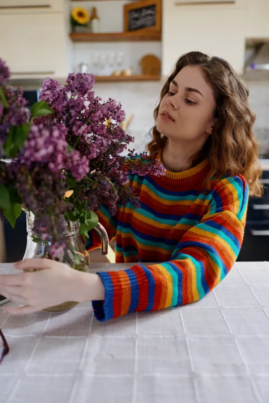 Woman lifestyle home, indoors, a woman arranges a bouquet of purple flowers on a table, wearing a colorful striped sweater, calm focused mood, bright kitchen background, cozy domestic scene.