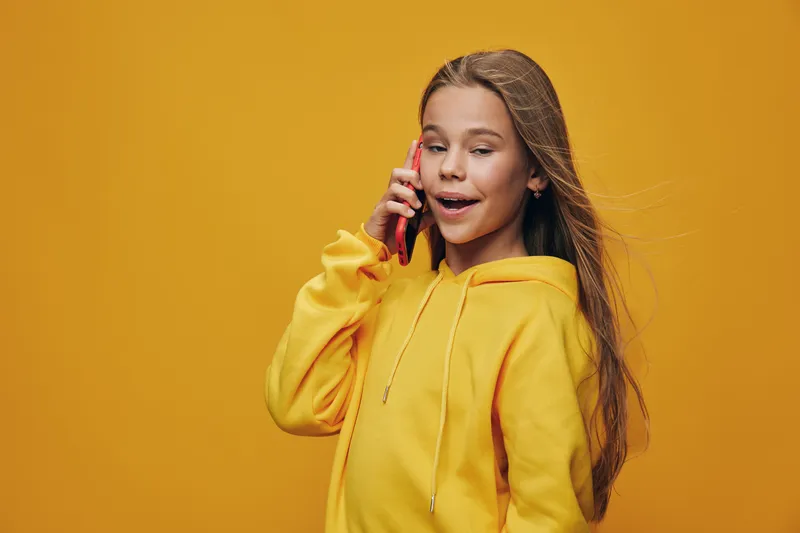 Girl in yellow hoodie talking on phone with happy expression on orange background. Long hair and casual outfit for communication and technology use by child.