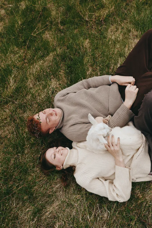 Young couple lies on grass holding a white baby goat together in cozy sweaters enjoying a peaceful outdoor moment in natural light and relaxed atmosphere.