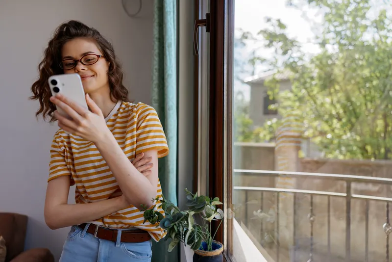 Cheerful woman indoors by a window, looking at smartphone with a smile, striped shirt, casual jeans, sunlit room, greenery nearby, relaxed lifestyle moment, natural colors, candid