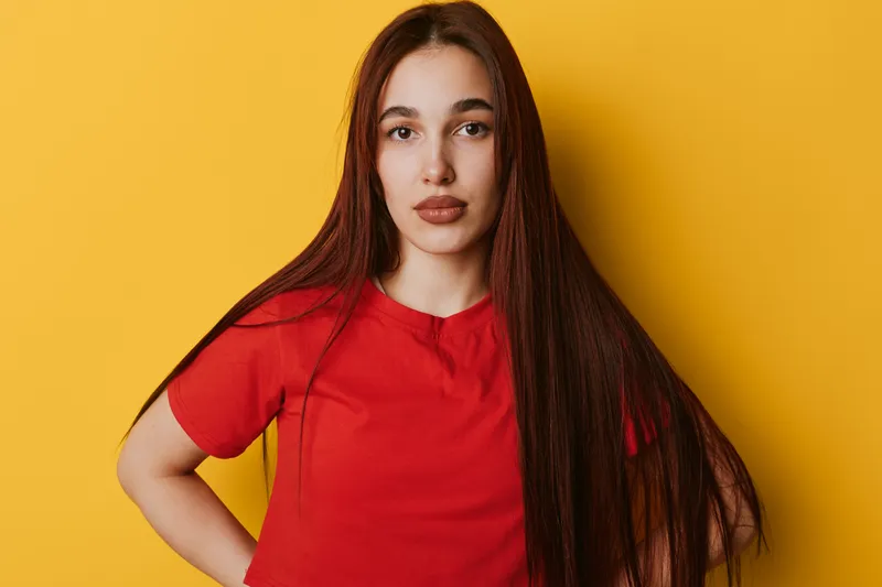 Young woman with long brown hair wearing red t shirt standing against yellow background looking at camera with confident expression and hand on hip.