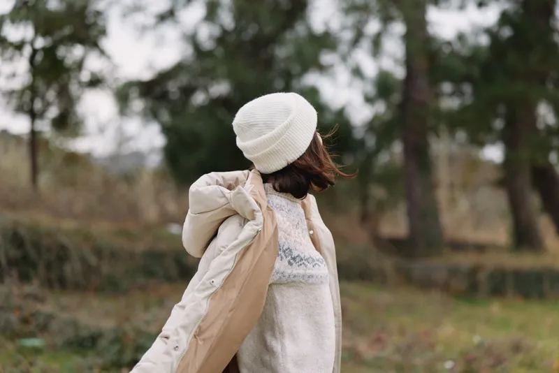 Woman in a beige coat and knit beanie standing in a park with autumn trees, looking away, soft natural light, casual winter fashion, cozy outdoor scene for lifestyle use.