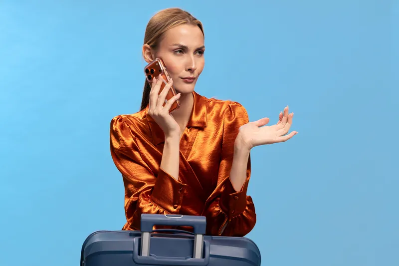 Blonde woman in shiny orange blouse holds smartphone near ear, gesturing with hand, sitting behind blue suitcase on light blue background. Concept of travel, communication, and planning.