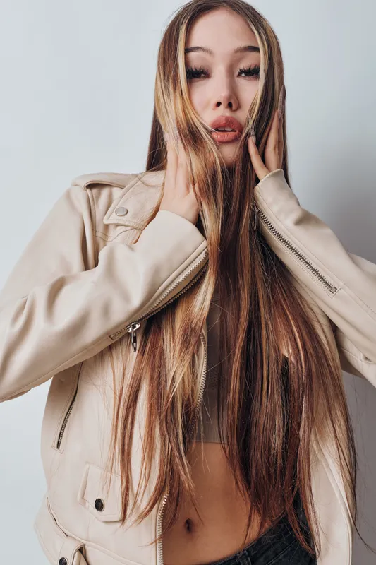 Young woman with long straight hair wearing light leather jacket posing against white background with hands near face, fashion portrait and confident expression.