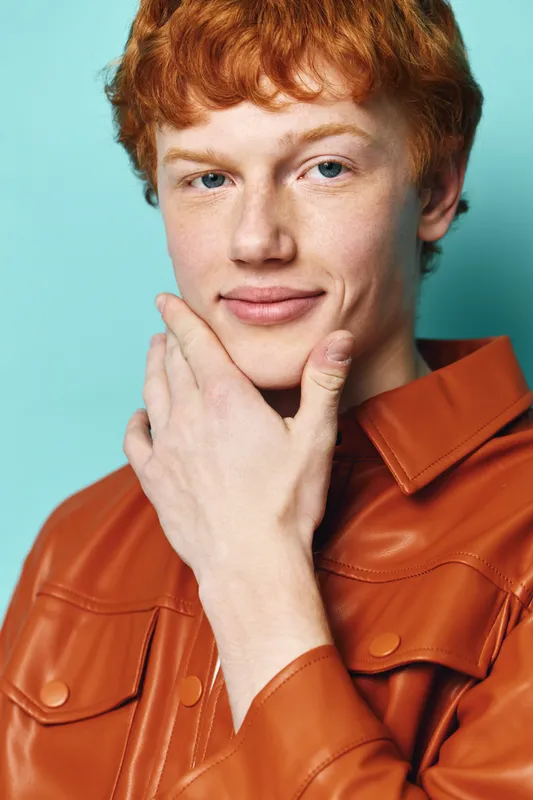 Young man with red hair and freckles wearing brown leather jacket touching chin looking at camera with confident expression on blue background in studio portrait closeup.