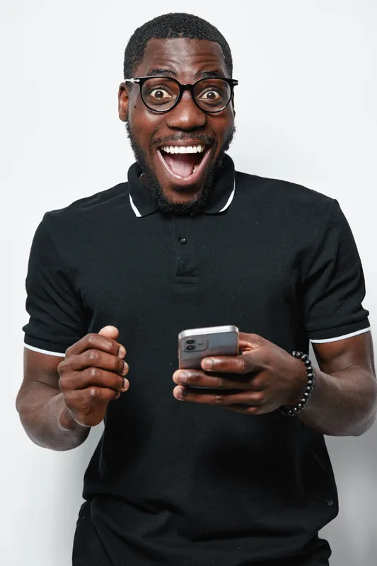 African American man with dark skin and glasses wearing black polo shirt holding smartphone with excited expression on white background. Technology, communication and happiness concept.