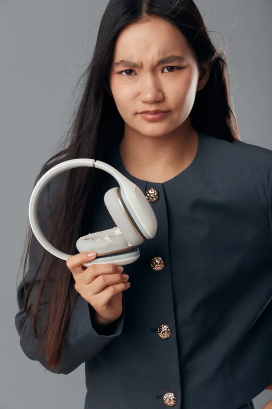Young woman with long dark hair holding white headphones and frowning, feeling displeased or confused, wearing a dark jacket, isolated on grey background with expression of dissatisfaction.