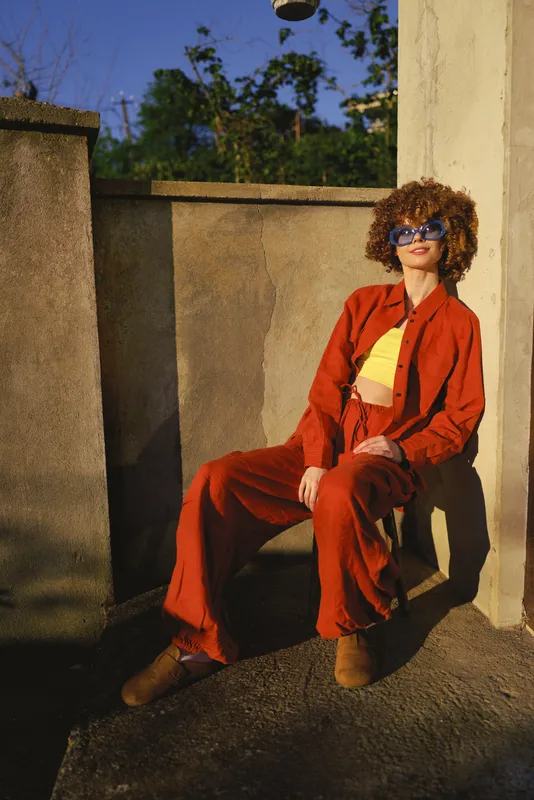 Young woman with curly hair sits on a sunlit rooftop ledge wearing a bold red suit and yellow top, smiling confidently in the warm urban light