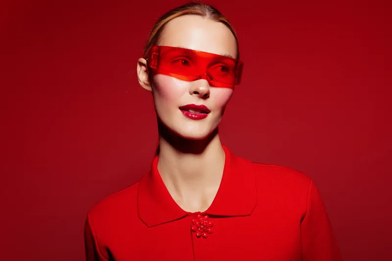 Young woman in red sweater and red translucent glasses poses against red background. Fashion portrait with bold color contrast and modern style. Concept of confidence and trend.