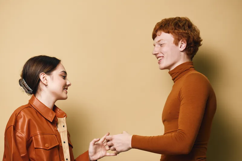 Couple in casual autumn fashion sharing a tender moment with warm smiles against beige background. Young man and woman dressed in brown and orange tones with natural expressions.