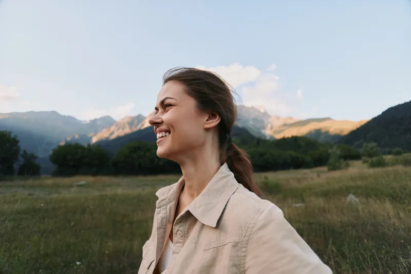 A smiling woman in a light beige jacket stands in a green meadow with mountains in the background, conveying fresh air, freedom, and outdoor lifestyle.