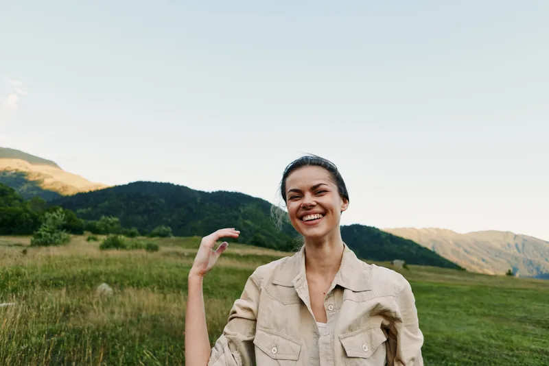 Young woman in a beige jacket laughs in a sunlit open field with distant mountains, capturing a carefree, natural lifestyle moment, a gentle breeze completes the scene.