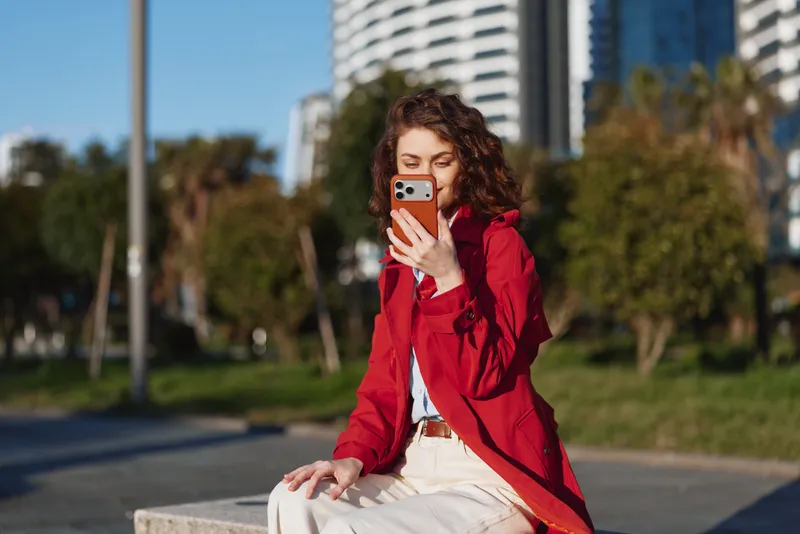 Smiling woman in a red jacket using a smartphone outdoors, seated on a bench. Urban park setting with tall buildings in the distance, casual lifestyle moment.