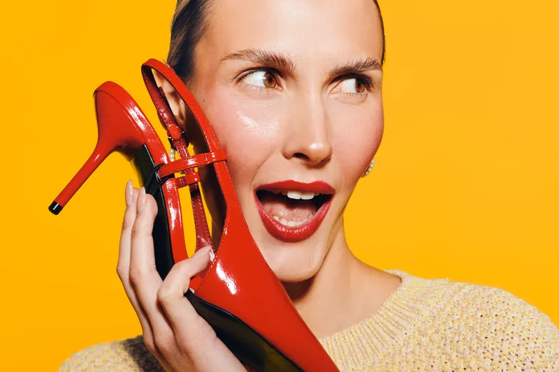 A cheerful woman with red lipstick holds a shiny red high heel shoe near her face against a bright yellow background. Concept of fashion, style, and vibrant colors.
