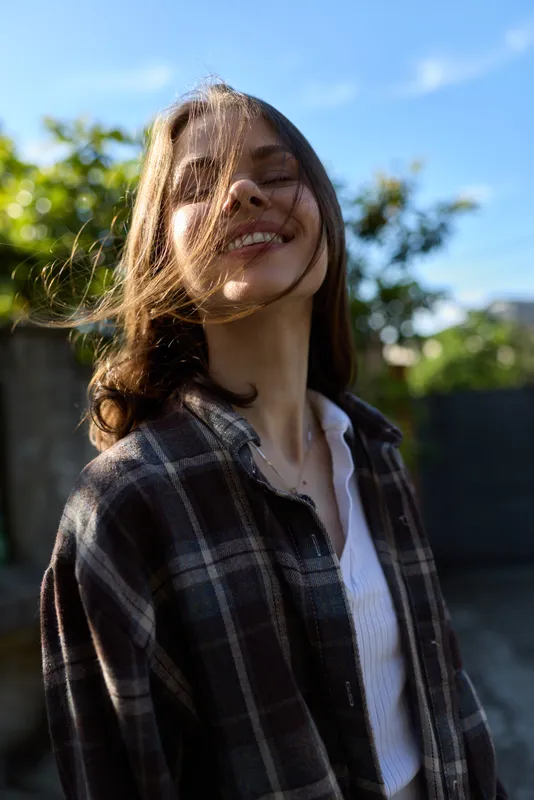 A young woman in a plaid shirt enjoys a sunny outdoor moment, hair swept by the breeze, smiling naturally in a candid lifestyle portrait