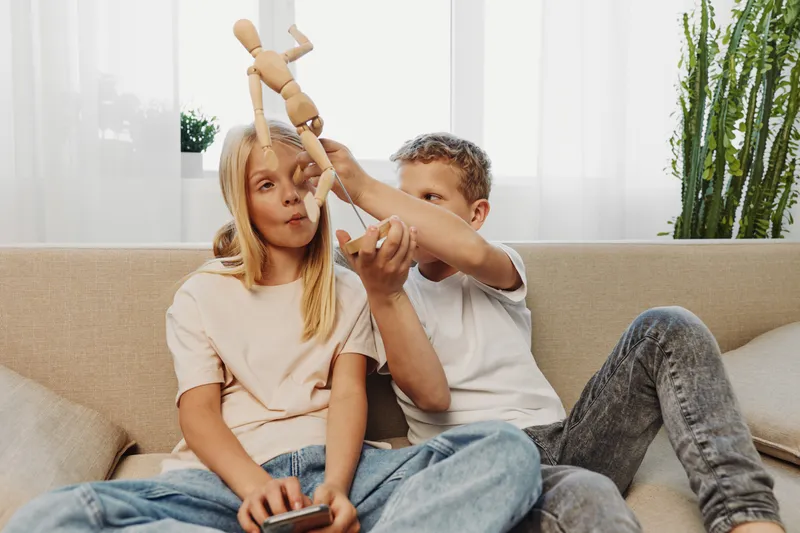 Two children sitting on sofa playing with wooden mannequin toy, enjoying leisure time in cozy home environment with natural light and indoor plants.