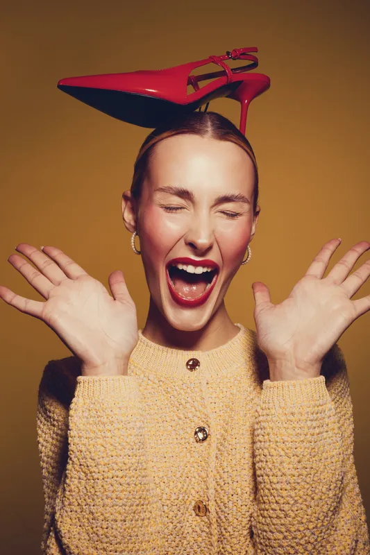 Happy woman with red high heel shoe on head laughs with eyes closed and hands raised. Studio portrait on yellow background. Concept of fashion, fun, and playful mood.