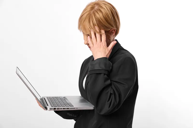 Stressed woman covers her face beside laptop in a bright white studio, worried expression, anxiety at work, coping with pressure concept