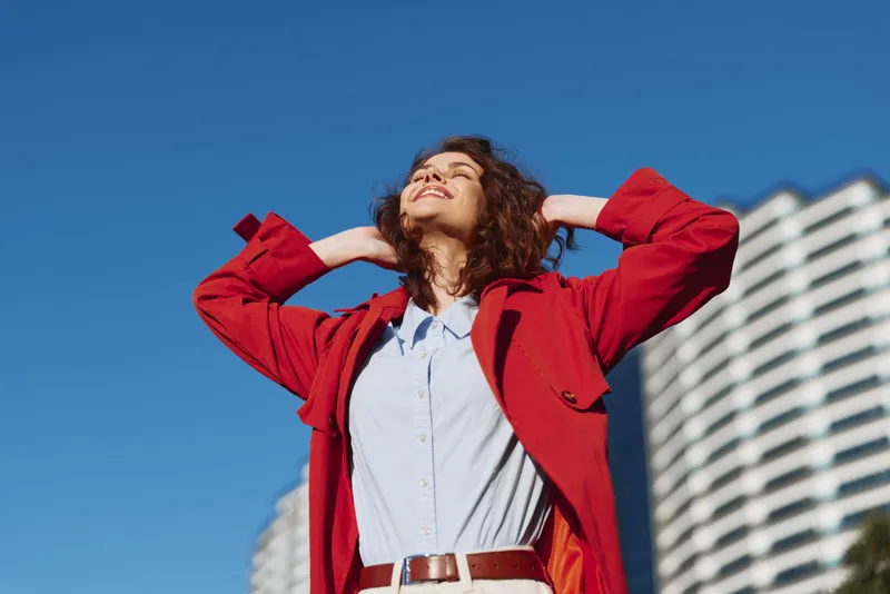 Young woman with curly hair enjoys a sunny day outdoors wearing a red jacket and white shirt with a joyful expression and relaxed posture.