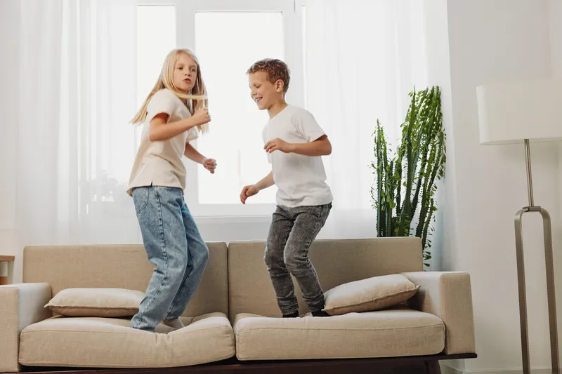 Happy children jumping on sofa in bright living room with large windows and plants. Casual home scene showing playful kids enjoying indoor activity and sibling fun with natural light atmosphere.