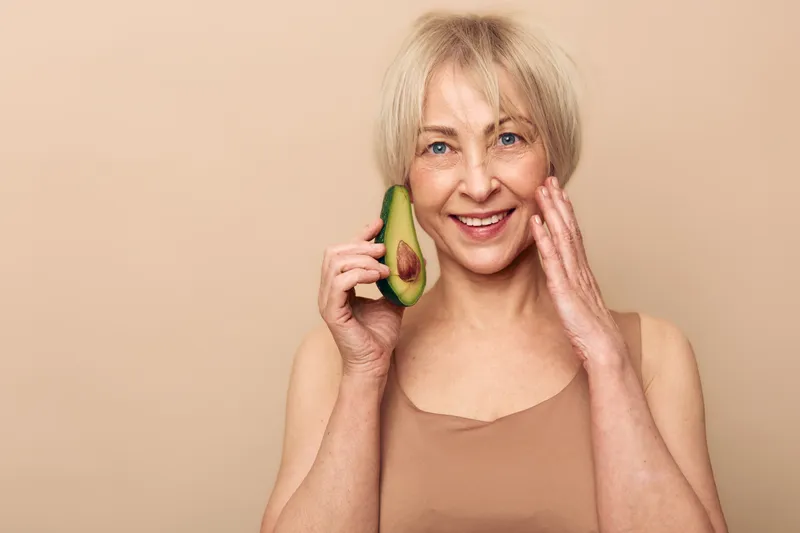 Happy mature woman with short blonde hair holds avocado half near face smiling gently on beige background. Concept for healthy lifestyle, natural beauty, skincare and wellness.