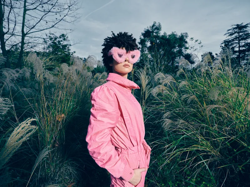Fashion portrait of a person in a pink jumpsuit wearing heart sunglasses outdoors among tall grasses, creating bold playful style with natural light and calm landscape.