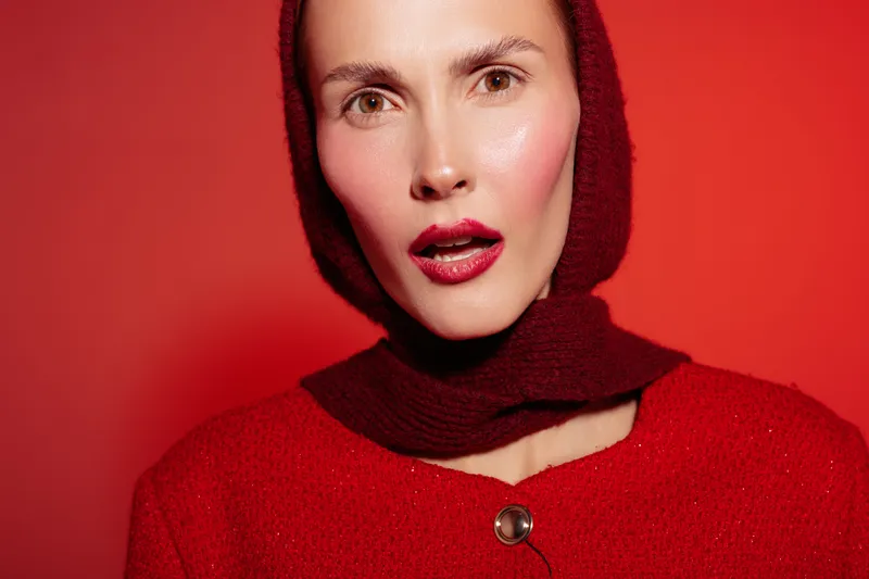Close-up portrait of a woman wearing a red scarf and red sweater against a red background. She has natural makeup and a confident expression. Fashion and beauty concept.